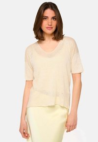 Femme aux cheveux bruns mi-longs portant un haut beige ample à col en V et à manches courtes, ainsi qu'une jupe jaune pâle, sur fond uni.