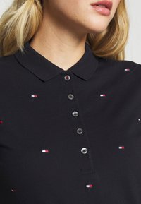 Svart pikétröja med krage, med små detaljer i röd, vit och blå färg samt grå knappar fram. Texturerat tyg.