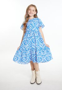 Mėlyna ir balta gėlių rašto suknelė, pagaminta iš lengvos medžiagos, su trumpomis rankovėmis, surinkta liemens dalimi ir plačia sijonu. Dėvima su smėlio spalvos batais.