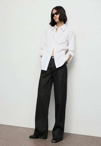 Camisa blanca de botón con mangas largas, combinada con pantalones vaqueros negros de pierna ancha y cintura alta. La modelo lleva gafas de sol negras y zapatos de tacón abiertos negros.