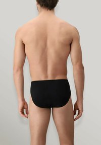 Slip da bagno neri realizzati in tessuto elasticizzato, con una texture liscia e una vestibilità aderente, posti su uno sfondo grigio chiaro.
