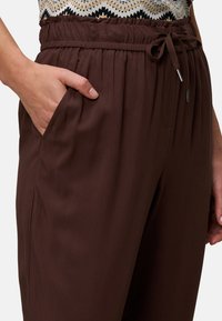 Hnědé lehké kalhoty s elastickým pasem, šňůrkou na stažení a bočními kapsami. Hladká textura a pohodlný volný střih.