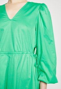 Vestido verde, leve, com decote em V, mangas bufantes, cintura elástica com cordão e textura suave e lisa.