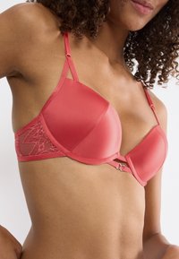 Różowokoralowy stanik z wyściółką, z koronkowymi bocznymi panelami, regulowanymi ramiączkami i centralnym zapięciem na hak. Gładka tekstura z subtelnym połyskiem.