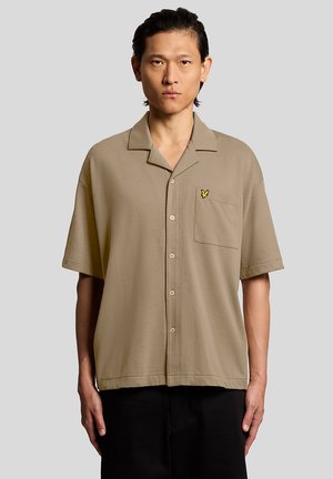 Homme aux cheveux foncés portant une chemise beige à manches courtes avec un petit logo jaune sur la poche, sur un fond uni.