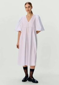 Weißes Baumwollkleid mit V-Ausschnitt, Puffärmeln und gerafftem Taillenteil. Das Modell trägt schwarze Slipper und transparente Knöchelsocken.