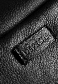 Fekete textúrájú bőr, rajta egy "DEPECHE" felirattal dombornyomott varrott címke. Az anyag pehelyszerű megjelenésű, enyhe fényességgel.
