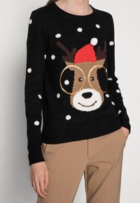 Pull noir à pois blancs avec un motif de chien de Noël portant de grandes lunettes, un chapeau de Père Noël rouge, et des accents de fourrure marron texturée.
