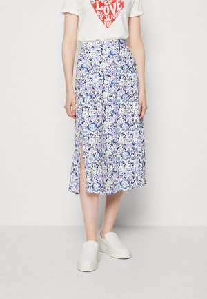 Femme portant un t-shirt blanc avec un texte cœur rouge et une jupe midi fleurie bleu et blanc avec une fente frontale, associée à des chaussures blanches sans lacets.