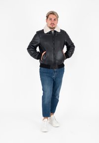 Giubbotto bomber in pelle nera con colletto in shearling bianco, chiusura con zip e polsini a costine. Indossato con jeans blu chiaro e sneaker bianche.