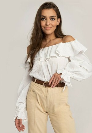 Blouse blanche à épaules dénudées avec un col et des manches volantés, associée à un pantalon beige taille haute et une ceinture marron avec une boucle en métal.