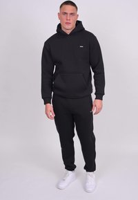 Un sweat à capuche noir et un jogging assorti en matériau doux, avec une poche frontale. Des baskets blanches apportent un contraste. Design simple avec un branding minimal.