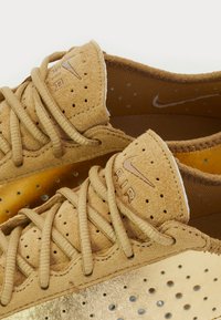 Kratak pregled bež suede Nike Air tenisica s tkanim vezicama i perforiranim zlatnim metalnim panelima na području prstiju.