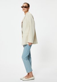 Blazer de gran tamaño en color beige claro, jeans de pierna recta azules y bailarinas blancas. Gafas de sol de montura redonda y pendientes tipo moño completan el look.