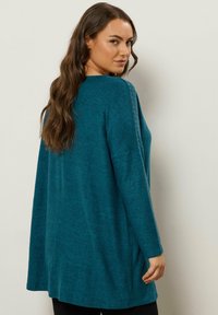 Maglione lungo oversize di colore teal con finitura texture. Presenta borchie lungo le cuciture delle spalle e una vestibilità rilassata.