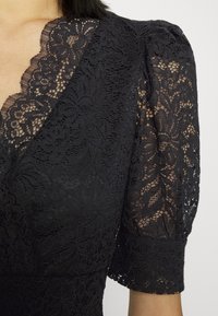 Schwarzes Spitzenkleid mit einem gewellten Ausschnitt und puffartigen Ärmeln, verziert mit floralen Mustern und transparenter Textur im gesamten Oberteil und an den Ärmeln.