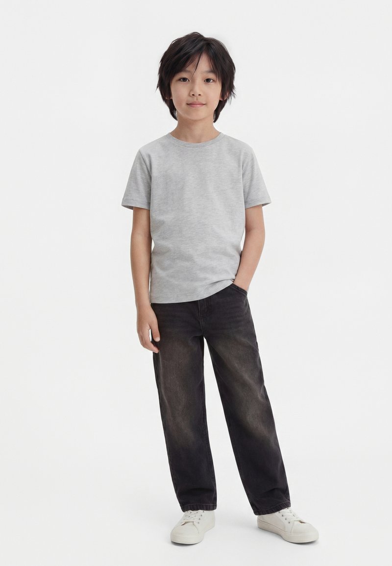 Garçon aux cheveux noirs portant un t-shirt gris clair, un jean noir ample et des baskets blanches, debout avec une main dans la poche sur un fond uni.