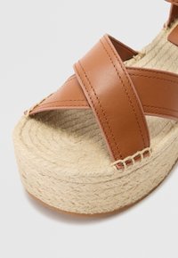 Sandales en cuir marron à plateforme avec des brides croisées, semelle en jute, et détails de couture texturée. La semelle a une forme ronde et plate.