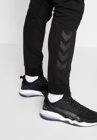 Svarta träningsbyxor med ett svart chevron-mönster på sidan, kombinerade med svarta sneakers med en vit sula och en strukturerad nätöverk.