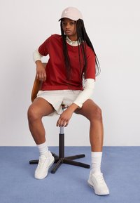 Röd kortärmad skjorta med vit krage och logotyp, i kombination med beige shorts. Vita sportskor och en ljusrosa keps. Sitter på en pall.