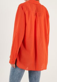 Femme portant une chemise orange vif à manches longues et ample, ainsi que des jeans bleus clairs, vue de dos contre un fond uni.