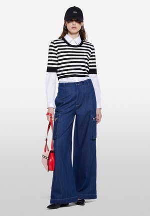 Maglione cropped a righe bianche e nere, camicia bianca sotto, pantaloni di denim blu larghi con tasche cargo, borsa rossa, cappellino nero e scarpe.