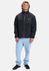 Giacca in fleece nera con colletto alto, chiusura con zip e tasca sul petto in tessuto grigio. Indossata sopra jeans blu chiaro e scarpe nere.