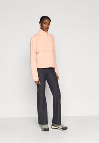 Ανοιχτό ροζ μπλουζάκι με φερμουάρ και μακριά μανίκια, με ψηλό γιακά, συνδυασμένο με σκούρο γκρι παντελόνι καμπάνα και γκρι αθλητικά παπούτσια με ροζ λεπτομέρειες.