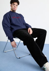 Sudadera de la marina con logo rojo, combinada con pantalones de pana negros, sentado en una silla plegable de metal sobre una alfombra azul. Estilo casual.