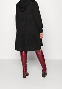 En person i en svart hoodieklänning står, iförd höga bourgogne stövlar med en kraftig klack, mot en ljusgrå bakgrund.