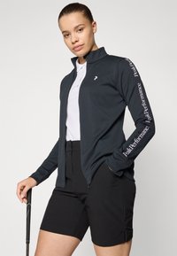 Marineblauwe ritsjack met hoge kraag, voorzien van een witte logo en 'Peak Performance' tekst op de mouw, gecombineerd met zwarte shorts en een golfclub.