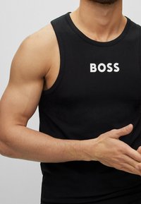 Muskulös person i svart linne med ordet "BOSS" i vita bokstäver, händerna placerade vid midjan mot en enfärgad bakgrund.