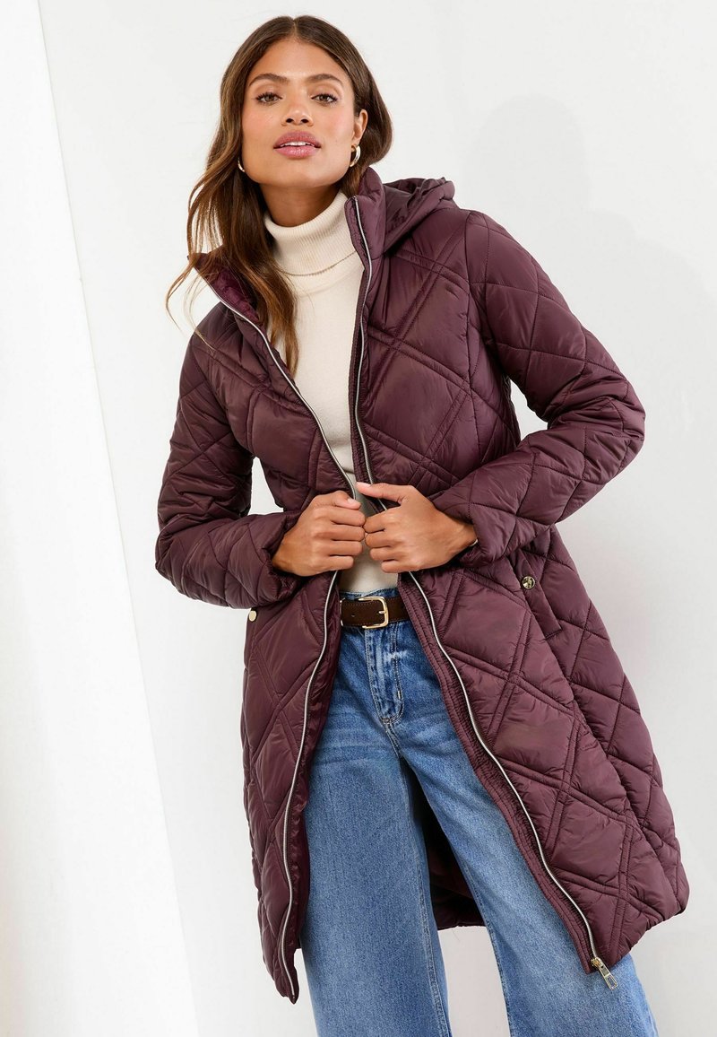 Vrouw in een open,gequilte marsala-jack met capuchon over een beige turtleneck, blauwe jeans en een bruine riem, staand tegen een witte achtergrond.