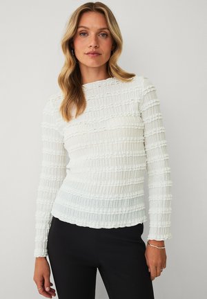 Kvinde med blondt hår iført en tekstureret hvid langærmet top og sorte bukser, stående mod en ensartet lys baggrund.