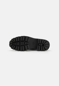 Semelle de chaussure en caoutchouc noir avec des rainures profondes et une surface texturée pour une meilleure adhérence. Accentuée par un logo près du talon et des sections surélevées pour un soutien supplémentaire.