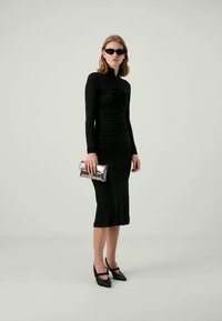 Schwarzes langärmeliges Kleid mit gerafftem Textil, knielang, kombiniert mit einer silbernen metallischen Clutch und schwarzen spitzen Absatzschuhen.
