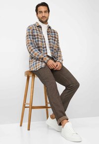 Geplooid overhemd in blauw, bruin en wit; gestructureerde stof. Bruine getailleerde broek; gladde afwerking. Witte sneakers. Houten kruk.