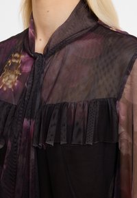 Nahaufnahme eines durchsichtigen schwarzen Tops mit Rüschen über einem floral gemusterten Stoff, getragen von einer Person mit heller Haut und blonden Haaren.