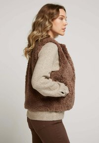 Gilet en fausse fourrure marron avec un col arrondi, des poches latérales et une texture douce, superposé à un haut beige clair à manches longues.