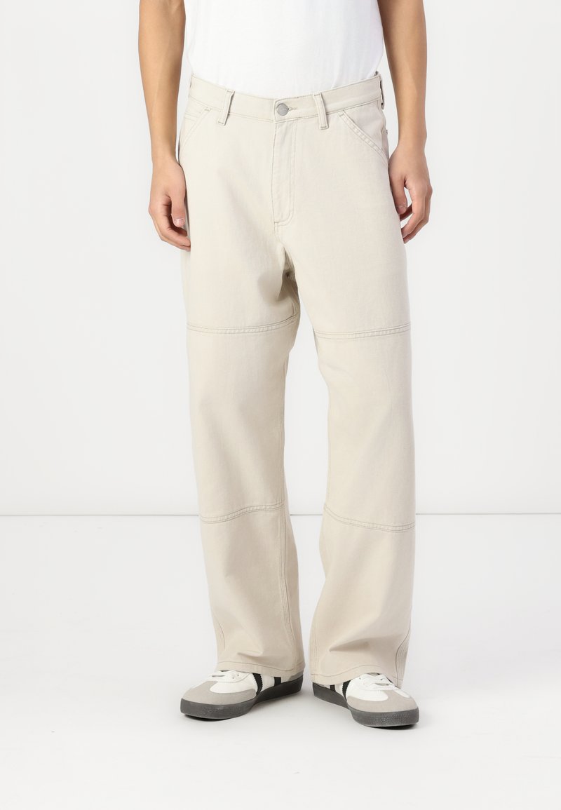 Beige wide-leg pants made of denim with contrasting stitching, front pockets, and a relaxed fit, paired with gray and white sneakers.