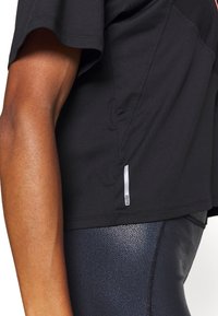 Camiseta atlética negra de manga corta con un panel superior texturizado, que presenta una etiqueta de logo reflectante en el lado y combinada con leggins negros brillantes.