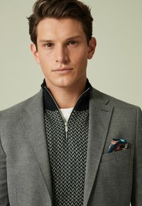 Grauer Blazer über einem gestrickten Pullover mit einem Reißverschlusskragen, der ein Zickzackmuster aufweist. Einstecktuch mit buntem Blumenmuster.