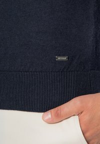 Dunkelblauer Pullover aus weichem Strickmaterial mit gerippten Bündchen und Saum. Besitzt ein kleines Metallschildchen in der Nähe der Naht.