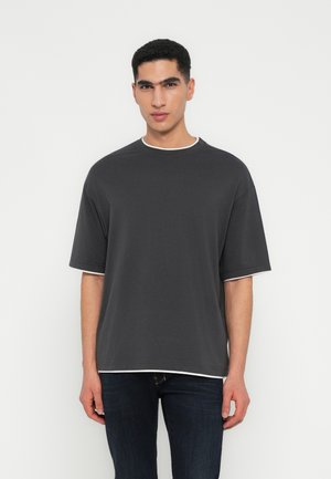 Jonge man in een donker oversized t-shirt over een wit shirt, gecombineerd met donkere jeans, staande tegen een effen witte achtergrond.