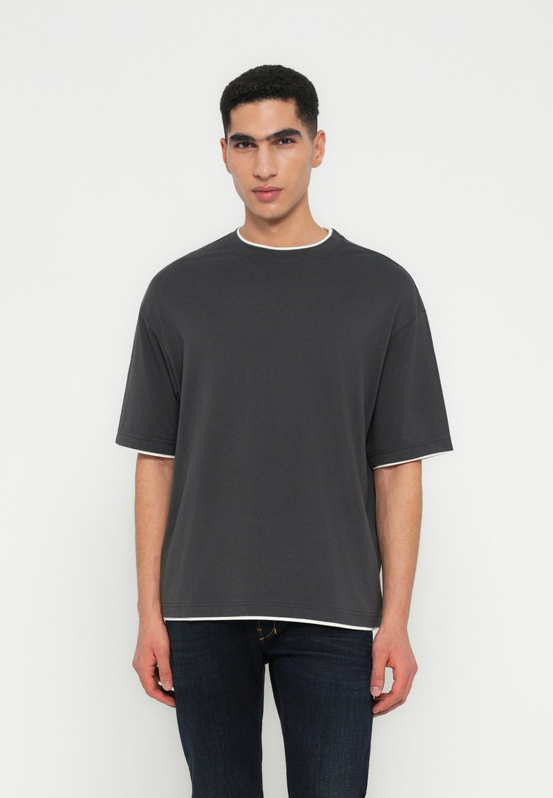 Jonge man in een donker oversized t-shirt over een wit shirt, gecombineerd met donkere jeans, staande tegen een effen witte achtergrond.