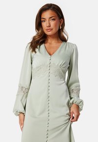 Zelené saténové šaty s dlouhými nafouknutými rukávy, krajkovými detaily v pase a na manžetách a ozdobnými knoflíky na přední straně.