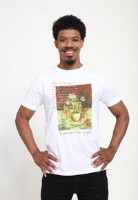 Bílá bavlněná trička s potiskem Van Goghova obrazu a červeným textem. Design zahrnuje květinovou vazbu v květináči. Natažené černé kalhoty.