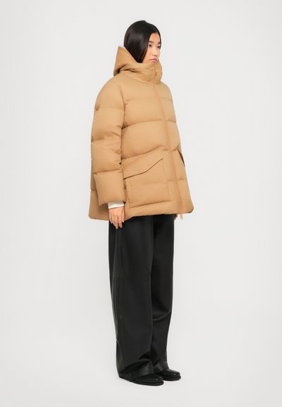 Eine Frau steht in einer beigen Steppjacke mit hohem Kragen, schwarzen weiten Hosen und schwarzen Schuhen vor einem schlichten hellen Hintergrund.