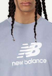 Világoskék pamut póló, prominent fehér "new balance" logóval és stilizált "N" mintával. Klasszikus kerek nyakú és rövid ujjú.