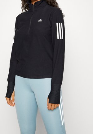 Kobieta ubrana w czarną długorękawową bluzę Adidas z suwakiem do połowy oraz jasnoniebieskie legginsy, obie części z białymi paskami Adidas na rękawach i nogawkach.